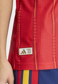 Red sports jersey with textured fabric, yellow accents, and a white Adidas logo label stating "AUTHENTIC LICENSED PRODUCT" near the hem.
