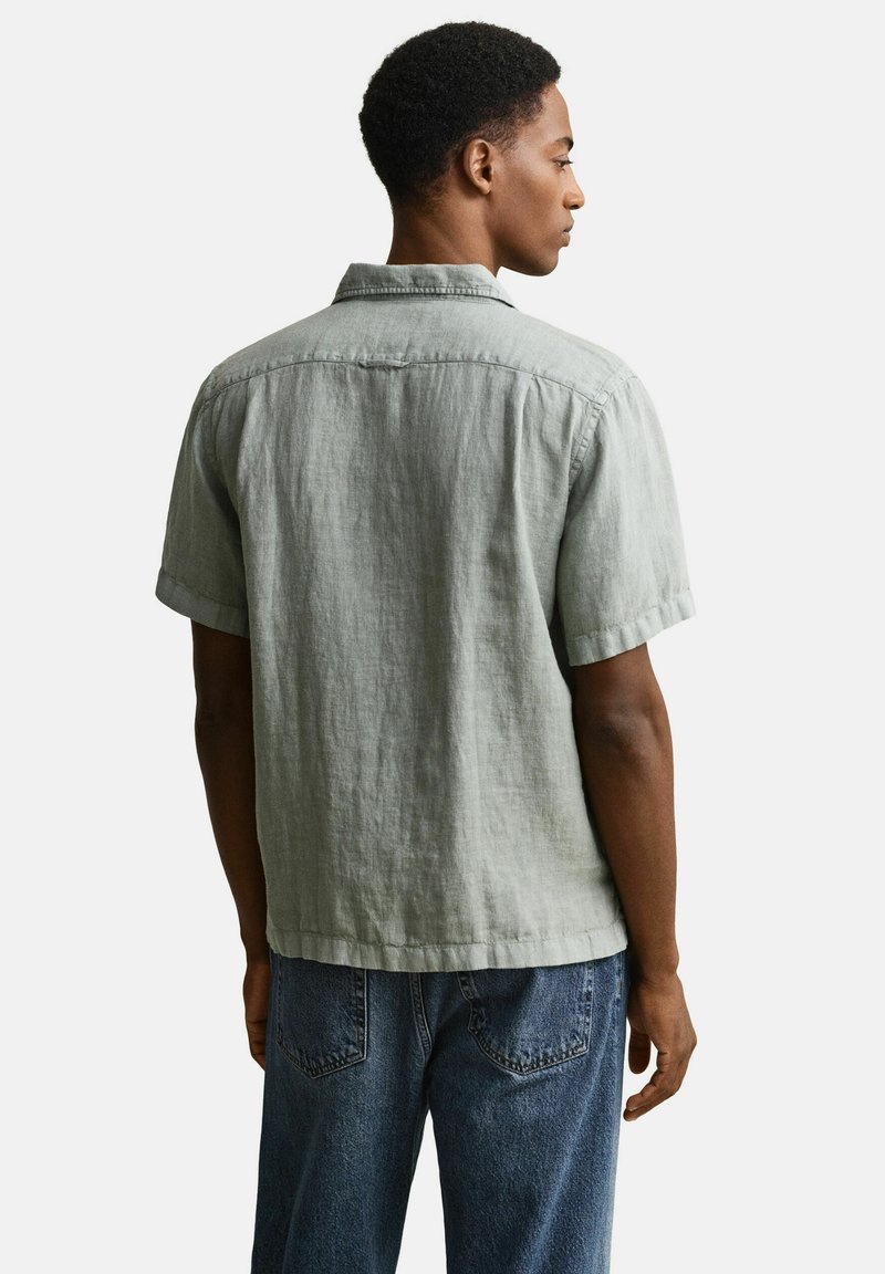 Uomo visto di spalle che indossa una camicia di lino a maniche corte color grigio chiaro e jeans di denim blu su uno sfondo bianco neutro.