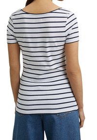 Vrouw die een wit T-shirt met korte mouwen draagt met zwarte horizontale strepen en een blauwe spijkerbroek, gezien vanaf de achterkant.