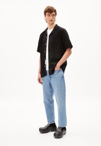 Zwarte korte mouw shirt, witte t-shirt eronder, lichtblauwe spijkerbroek en zwarte klompen. Het shirt heeft een zak, een casual pasvorm en een zachte textuur.