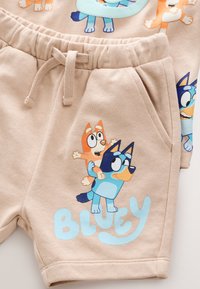 Shorts beiges avec des personnages de dessin animé en couleurs vives, taille à cordon de serrage et poches latérales. Texte bleu "BLUEY" visible.