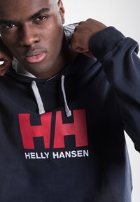Sudadera de color azul marino con cordones grises, que presenta un destacado logo de Helly Hansen en rojo y blanco. El material parece suave y con textura.