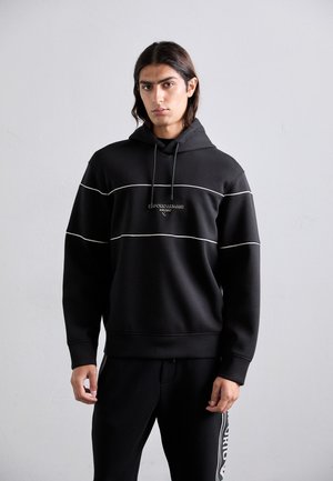 Ung mann med skulderlangt mørkt hår, iført svart hettegenser og svarte bukser med side/logo-striper, som står mot en grå vegg.