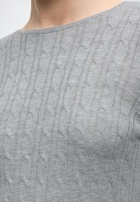 Gros plan d'une personne portant un pull en maille torsadée gris clair avec un col rond.