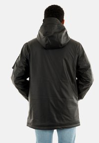 Giacca nera impermeabile con cappuccio, caratterizzata da una texture liscia, maniche lunghe e un orlo leggermente curvato. Vista posteriore.