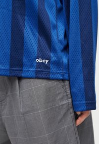 Μπλε και ναυτικό ριγέ μακρυμάνικο πουκάμισο από διχτυωτό ύφασμα με το λογότυπο "obey" πάνω από γκρι καρό παντελόνι, το χέρι ακουμπάει στο πλάι.
