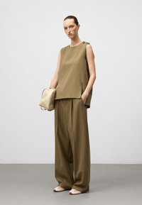 Kvinna som bär en lös olivgrön ärmlös topp och vida byxor, håller en beige liten ryggsäck och står framför en enhetlig bakgrund.