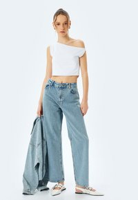 Top court blanc à épaules dénudées, jeans taille haute bleu clair, tenant une veste en denim ; mules blanches à bout pointu, décontracté, coupe ample.