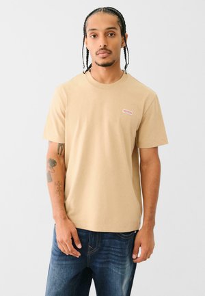 Beige Baumwoll-T-Shirt mit kurzen Ärmeln, rundem Halsausschnitt und einem kleinen roten und weißen Markenlabel auf der linken Brust. Getragen mit blauen Jeans.