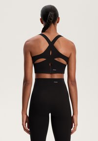 Črn športni modrček s križnimi naramnicami in izrezki, v kombinaciji z visokimi črnimi legicami. Narejeno iz raztegljivega materiala, logotip blagovne znamke je viden.