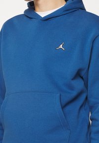 Sudadera azul hecha de una tela suave con un bolsillo frontal. Presenta un pequeño logo en blanco y negro de Jumpman en el pecho. Diseño simple y funcional.