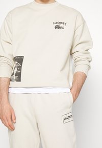 Personne portant un sweat-shirt beige Lacoste et un pantalon assorti avec des logos et textes noirs, un sous-vêtement blanc visible à la taille.