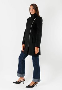Cappotto nero con colletto alto, zip asimmetrica e vestibilità strutturata. Indossato sopra jeans in denim scuri e tacchi a punta neri.