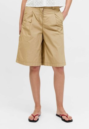 Persona che indossa pantaloncini beige fino al ginocchio, top bianco con occhielli e infradito rosse, in piedi con le mani in tasca su sfondo semplice.