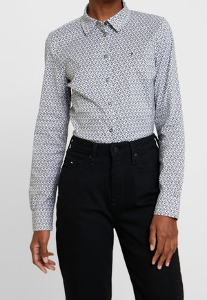 Gros plan sur une personne portant une chemise blanche à manches longues avec des motifs, boutonnée et rentrée dans un jean noir taille haute.