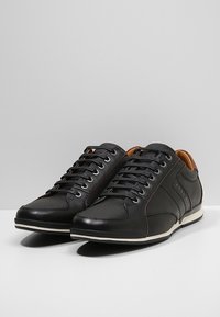 Baskets noires avec une tige texturée, des accents en cuir lisse, des lacets marron et une semelle en caoutchouc blanche. Présentent un motif cousu sur les côtés.