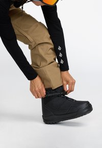Botte de neige noire avec une surface texturée, maintenue par une sangle grise. Pantalon beige avec une texture lisse, arborant un détail logo sur la manche.
