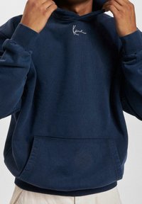 Persona ajustando la capucha de una sudadera azul marino con un pequeño logo bordado en blanco en el pecho y un bolsillo delantero.