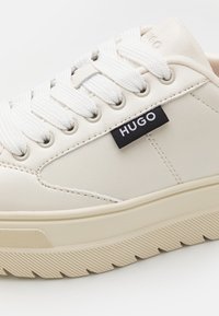 Zapatilla blanca de cuero con acabado texturizado, cordones redondos y un destacado logo negro "HUGO" en el lateral. Presenta una suela acanalada de color crema.