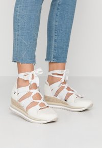 Fötter som bär vita kilklackssneakers med ankelsnören, tillsammans med ljusblå avklippta jeans mot en enfärgad bakgrund.