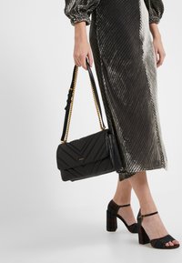 Czarna pikowana skórzana torebka na złotym łańcuszku, z klapką i wytłoczonym logo. Dopasowana do metalicznej, paskowanej spódnicy i wzorzystych szpilek.