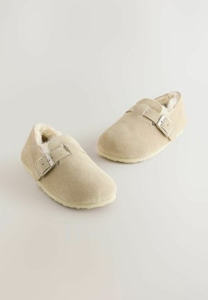 Beżowe zamszowe klapki z miękkim sztucznym futrem w środku, z metalowym paskiem z klamrą oraz teksturowaną gumową podeszwą. Okrągły kształt noska.