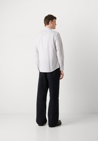 Camisa blanca ligeramente rayada con cuello abotonado y mangas largas, combinada con pantalones oscuros de pierna ancha y zapatos negros.