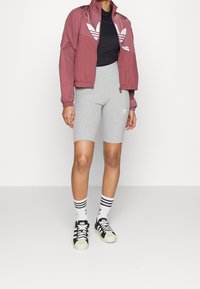 Vínová bunda na zip s bílým logem, černý přiléhavý top, šedé cyklistické šortky, černobílé pruhované ponožky a černé tenisky s bílými detaily.