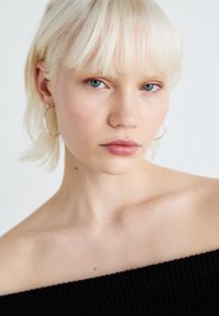 Modella con capelli corti biondo platino, che indossa orecchini a cerchio, top nero con spalle scoperte, pelle liscia, trucco neutro, occhi blu.