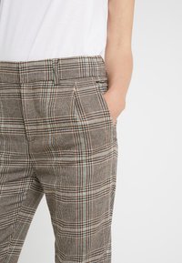 Spodnie w kratę w kolorach brązowym, beżowym i morskim o dopasowanym kroju. Posiadają dwie boczne kieszenie oraz plisowany przód.