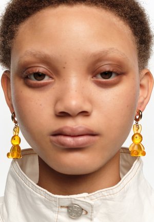 Zbliżenie osoby noszącej przezroczyste żółte kolczyki w kształcie króliczków oraz jasną koszulę z wysokim kołnierzem na białym tle.