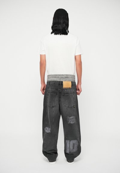 Jean en denim noir large avec une taille décolorée, plusieurs poches et des motifs imprimés incluant un logo et des chiffres. Texture douce.