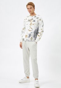 Bluza tie-dye w kolorach białym, szarym i beżowym; zestawiona z jasnoszarymi spodniami dresowymi i białymi trampkami. Gładki materiał, luźny krój, ściągacze na rękawach.