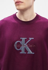 Bordový sweatshirt vyrobený z bavlny, s kulatým výstřihem, vyšívaným logem "Calvin Klein" v kontrastních barvách a volným střihem.