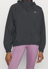 Černá větrovka s vysokým límcem, kapucí, elastickými manžetami a stahovacím pasem. Na hrudi má malé bílé logo.