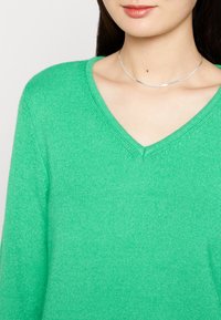 Green V-neck sweater made of soft, textured fabric. Features long sleeves and a subtle knit pattern, paired with a thin silver necklace.