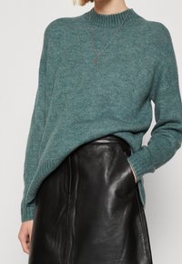 Pull en maille teal avec un col rond côtelé et des épaules tombantes, associé à une jupe en cuir noir au toucher lisse.