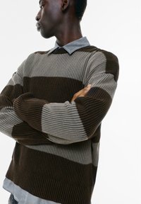 Sweter dzianinowy w poziome paski w ciemnym brązie, jasnym brązie i szarości, z prążkowaną teksturą oraz szarym podkoszulkiem z kołnierzykiem pod spodem.