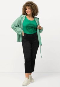 Sudadera verde con cremallera sobre una camiseta sin mangas verde con un diseño floral negro, combinada con pantalones cortos negros y zapatillas blancas.