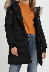 Musta toppatakki vetoketjulla edessä, vaakasuorilla tikatuilla saumoilla, hupulla jossa keinoturkiskoriste ja resorireunuksilla hihoissa. Päällä oranssista neuloksesta valmistettu teksturoitu neule.