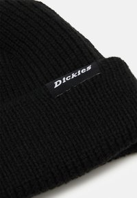 Črna pletena kapa iz teksturirane tkanine, ki ima črn značko z belim besedilom. Preprost, funkcionalen dizajn z ovitkom.