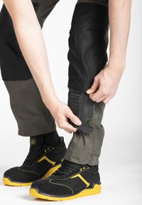 Pantalon de travail noir et olive avec des bretelles réglables ; tissu texturé ; une personne en train de fermer une fermeture ; chaussures noires avec des accents jaunes.