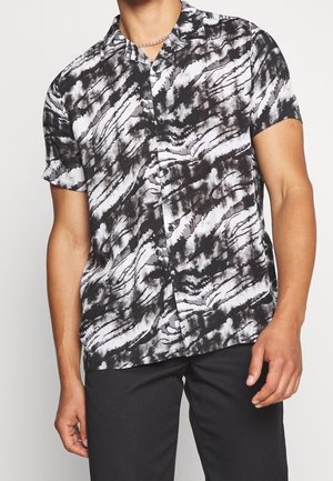 Man met een korte mouwen overhemd met zwart-wit abstracte tie-dye patroon en zwarte broek tegen een effen achtergrond.