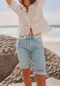 Blusa blanca de encaje con detalles bordados y un lazo en la parte delantera, combinada con pantalones cortos de mezclilla azul claro con una rodilla desgastada. Fondo oceánico.