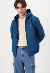 Blå huva zip-jacka med strömlinjeformat design, featuring en liten gul logotyp på bröstet, parat med en ljusbeige skjorta och denimjeans.