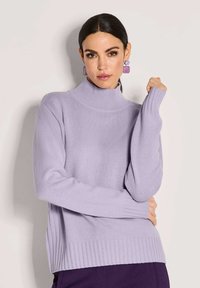 Eine Frau mit dunklem Haar trägt einen lavendelfarbenen Rollkragenpullover und passende geometrische Ohrringe und posiert vor einem schlichten hellen Hintergrund.