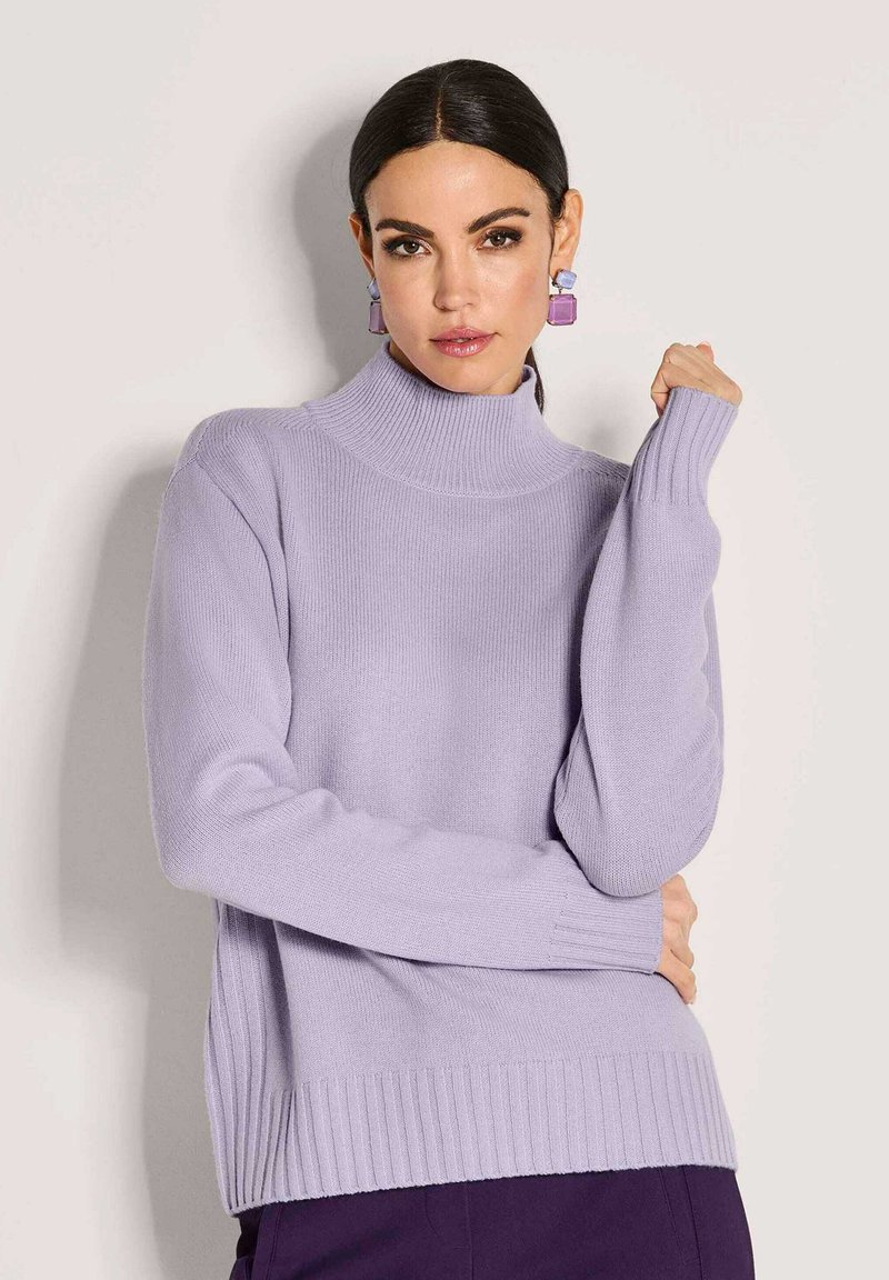 Eine Frau mit dunklem Haar trägt einen lavendelfarbenen Rollkragenpullover und passende geometrische Ohrringe und posiert vor einem schlichten hellen Hintergrund.