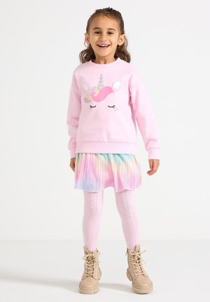 Jasnoróżowa bluza z kapturem z wzorem jednorożca, plisowana spódnica w tęczowe pasy, różowe rajstopy z wzorem oraz beżowe botki na masywnym obcasie.