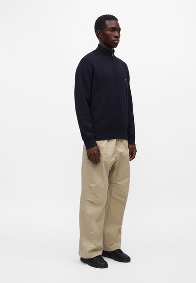 Homme debout portant une veste noire zippée, un pantalon beige à jambes larges et des chaussures noires sur un fond blanc uni.