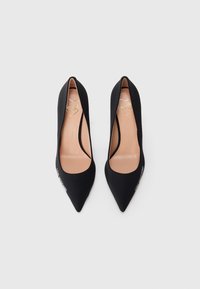 Pantofi cu toc înalt, vârful ascuțit, de culoare neagră, cu o textură fină, având o tăietură adâncă pe laterale și o branț de culoare bej cu branding auriu.